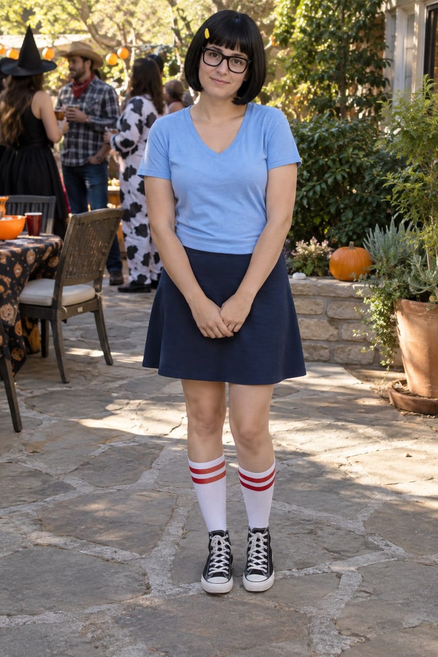 An adult dressed as Tina Belcher in a light-blue tee, navy skirt, glasses and canvas sneakers on a sunny patio.