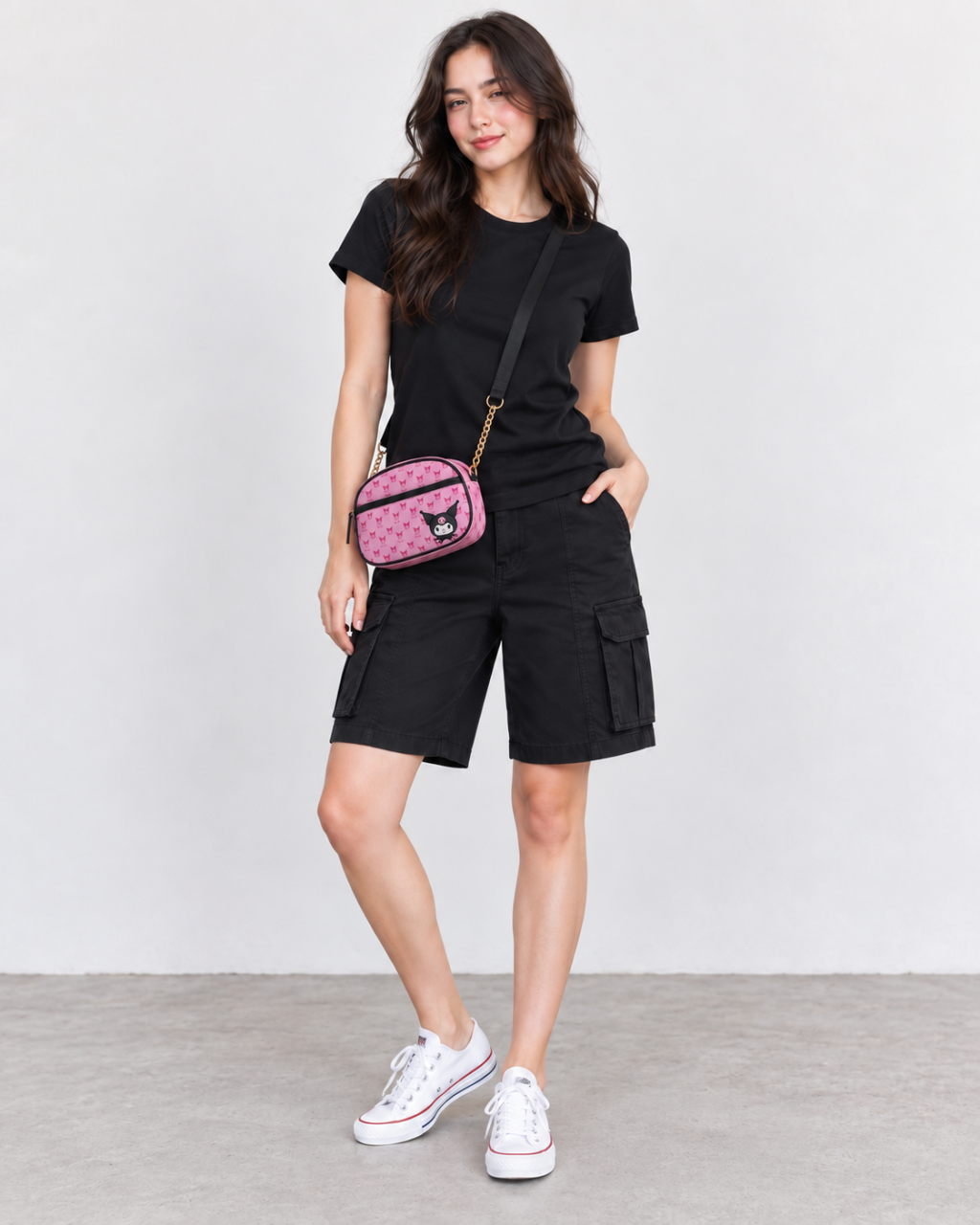 Adult woman wearing a black crew tee, black cargo Bermuda shorts, white canvas sneakers and a pink crossbody printed with repeating Kuromi skull motifs.